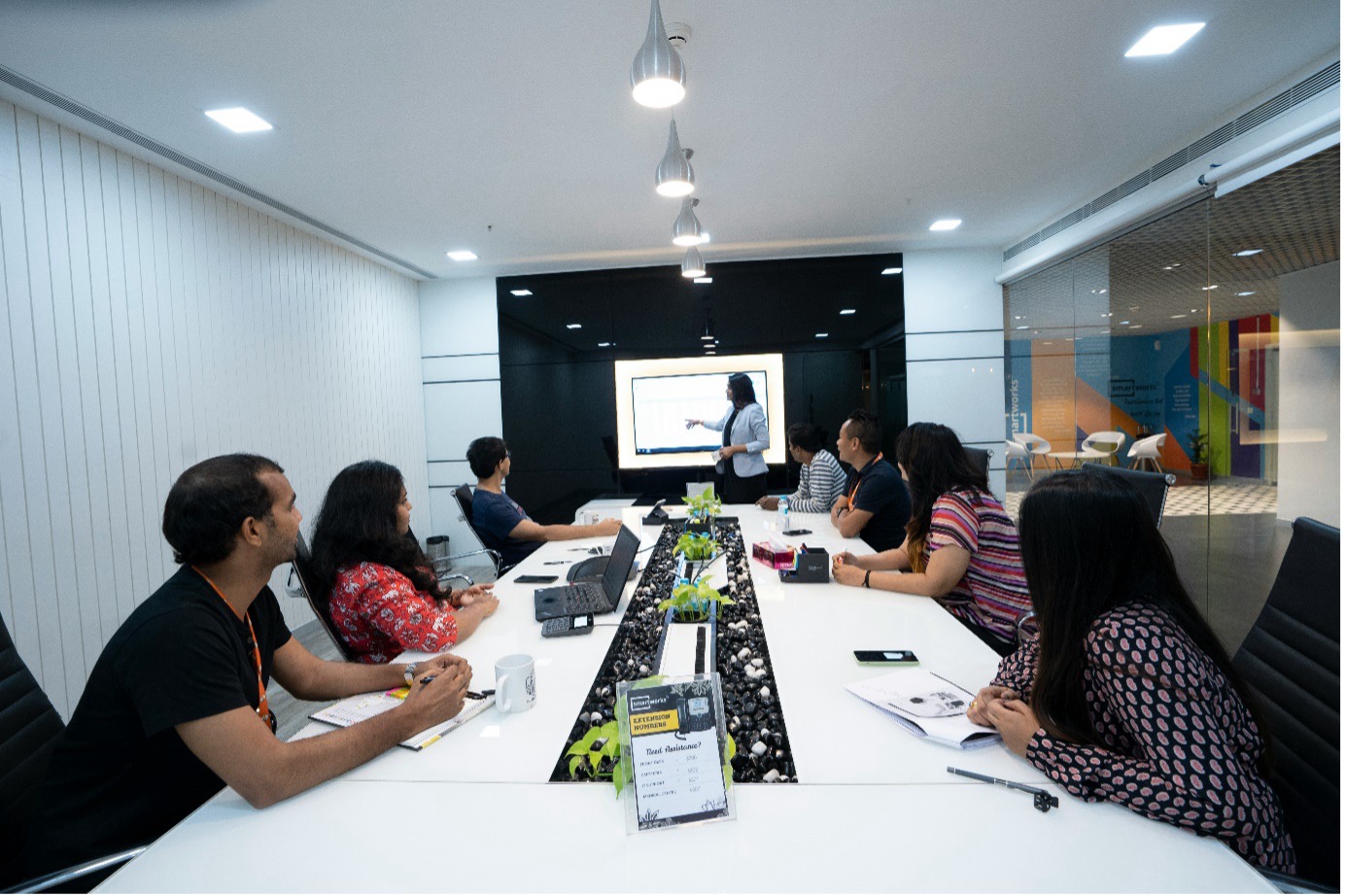 Professional photograph of a team sitting along a long conference table in a modern boardroom while a presenter at the front display outlines project scope and milestones using a Statement of Work template.