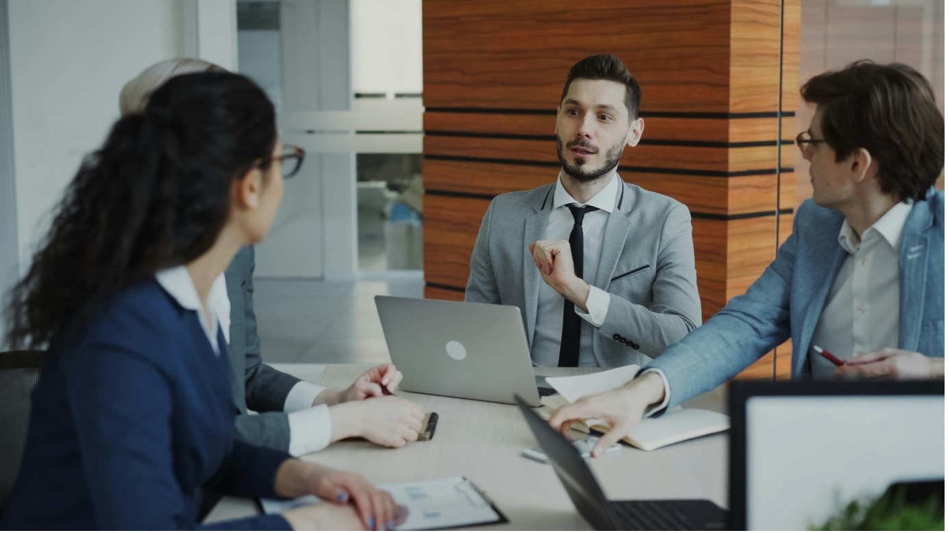 Photograph of corporate professionals in suits discussing project governance and change control structures around a conference table with laptops and documents, using a Statement of Work template.
