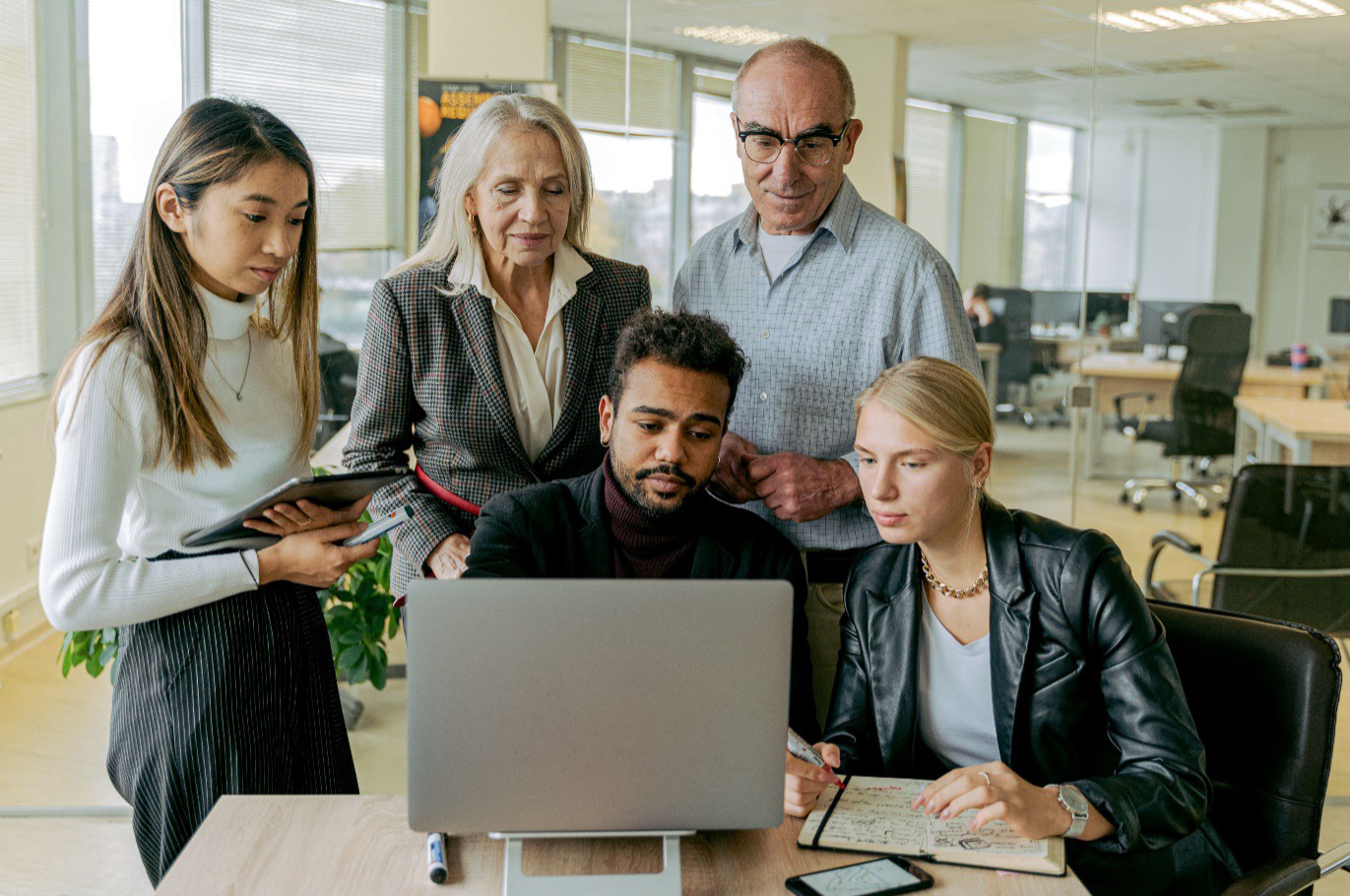 A multi-generational corporate team gathered around a laptop, exemplifying inclusive candidate selection by technology executive search firms.