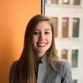 Jeune femme souriante avec de longs cheveux bruns, portant une chemise grise, devant un mur orange.
