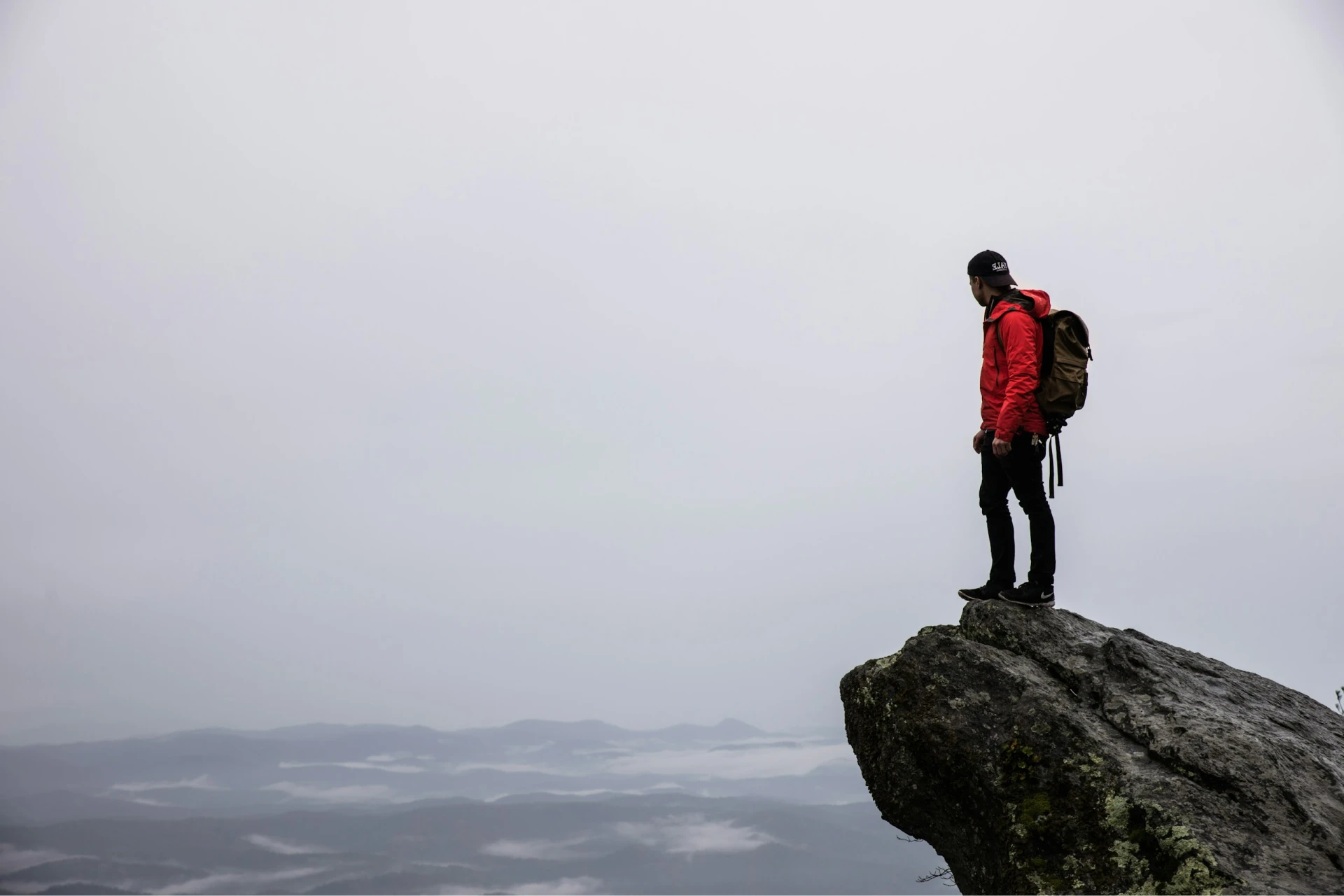 Homme en veste rouge et sac à dos debout sur un rocher regardant une vallée brumeuse.