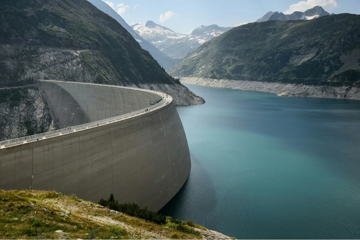 Barrage en béton incurvé retenant un lac entouré de montagnes avec des sommets enneigés.