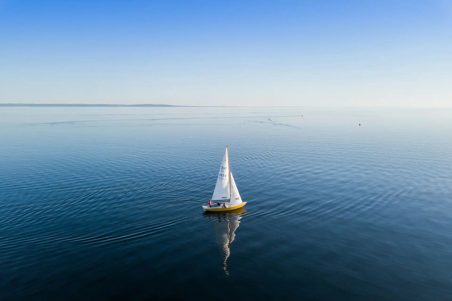 Voilier jaune avec voile blanche naviguant sur une mer calme et réfléchissante par temps clair.