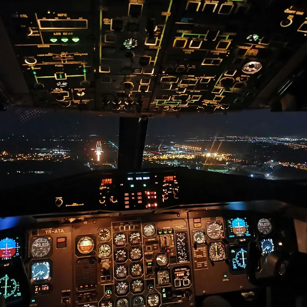Poste de pilotage d'un avion atterrissant de nuit vers une piste illuminée dans une ville.