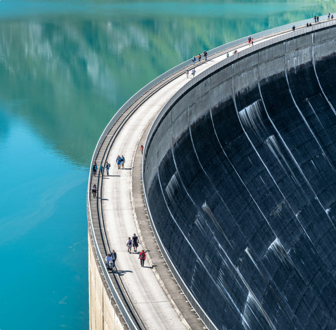 Des gens marchent sur le sommet d'un barrage en béton incurvé au-dessus d'un lac bleu.