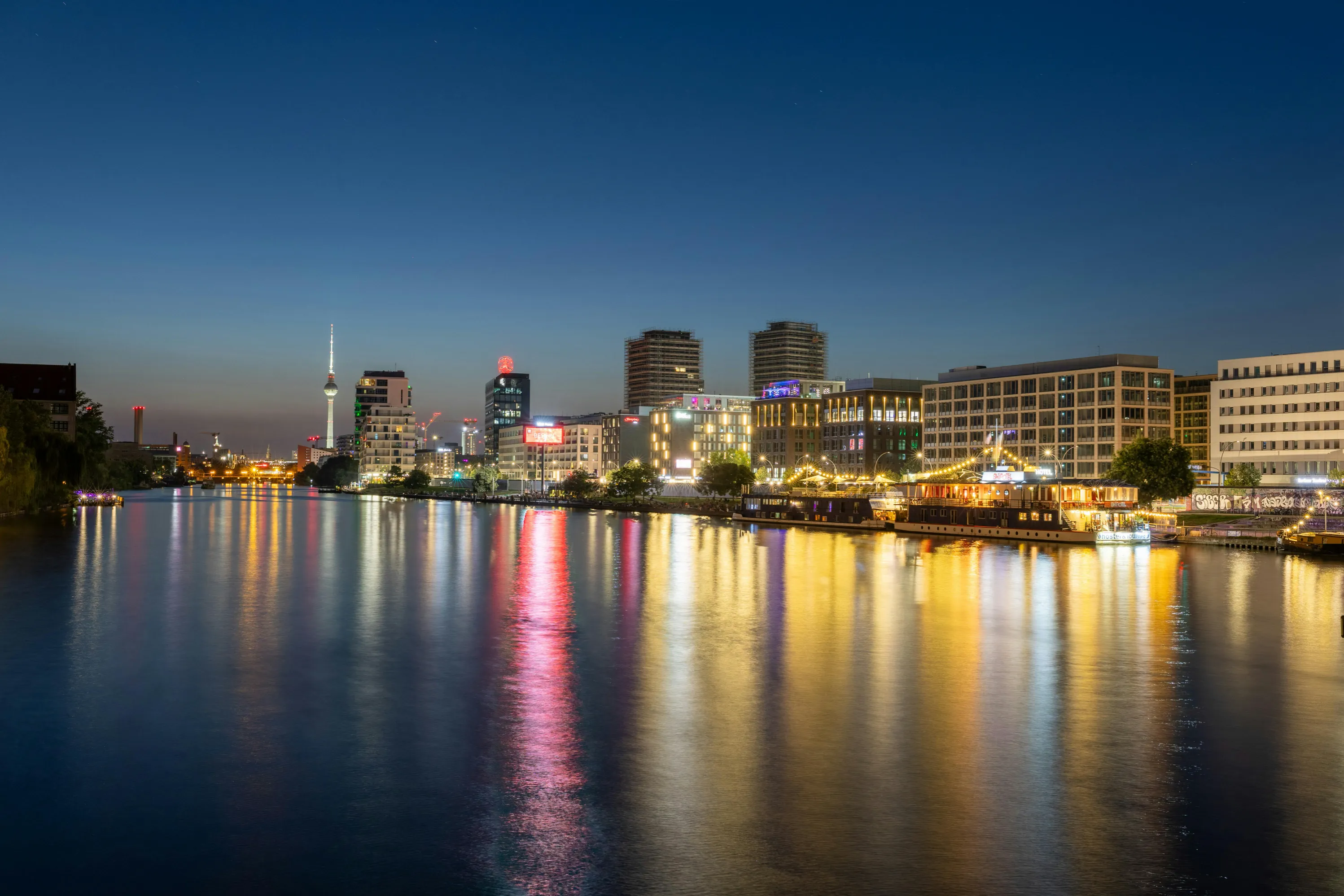 Abendlicher Blick auf die Berliner Skyline an der Spree mit beleuchteten Gebäuden und Spiegelungen im Wasser.