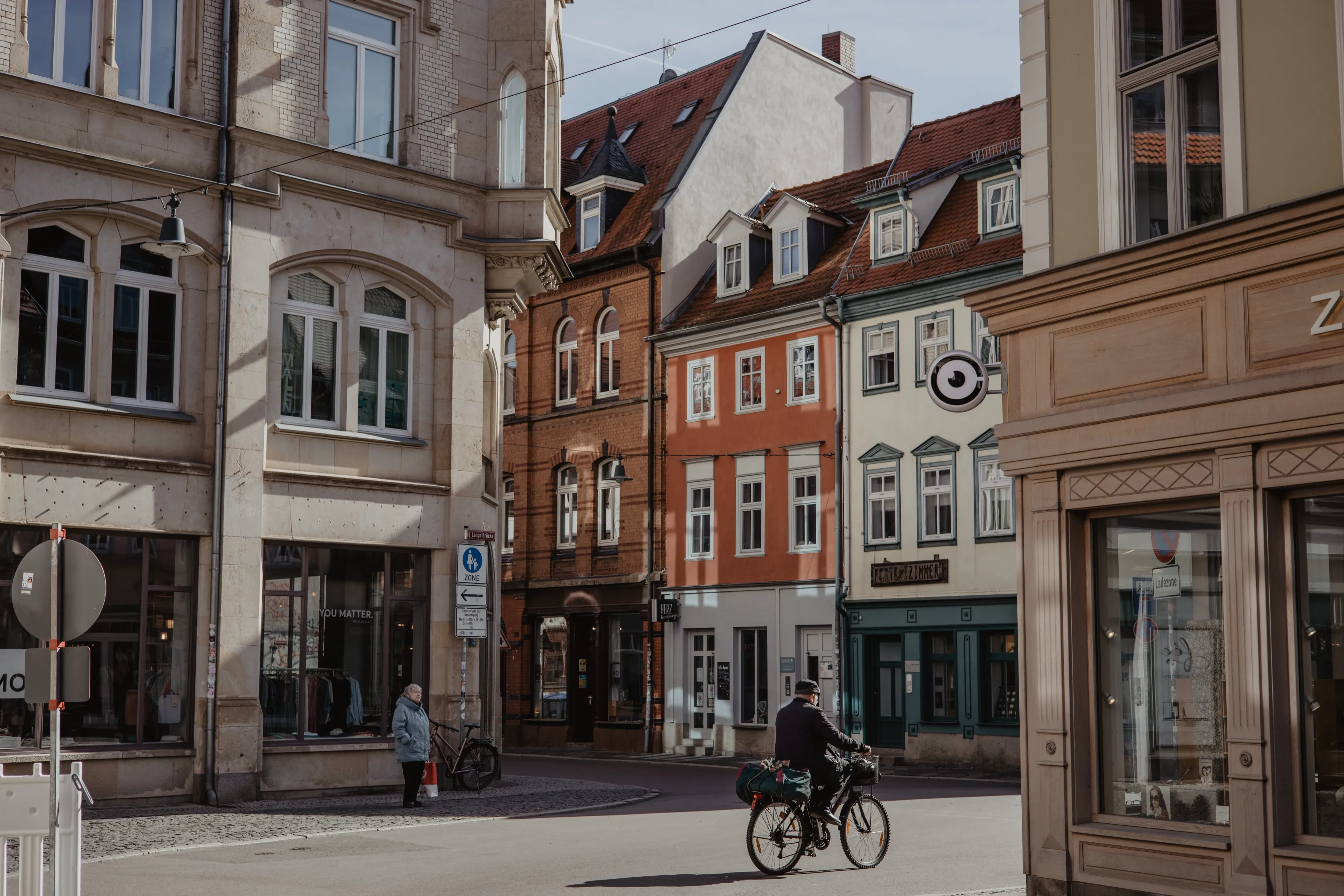 Straßenszene in der Osnabrücker Altstadt mit historischen, farbenfrohen Gebäuden und roten Ziegeldächern. Ein Radfahrer fährt über eine ruhige Kreuzung, während Passanten vor den Geschäften stehen. Die Fassaden aus verschiedenen Epochen verleihen der Straße einen charmanten, historischen Charakter.