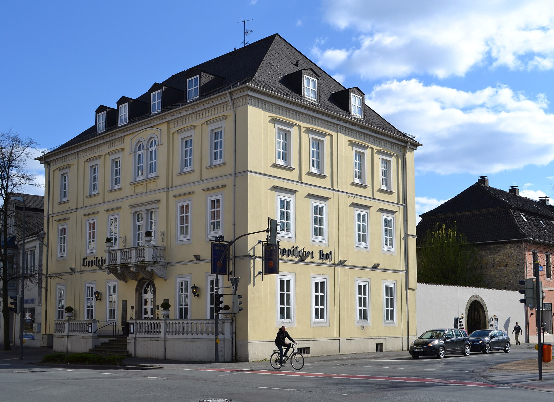 „Elegantes Hotelgebäude in München an einer von Bäumen gesäumten Straße, Auto fährt vor dem Eingang vor."
