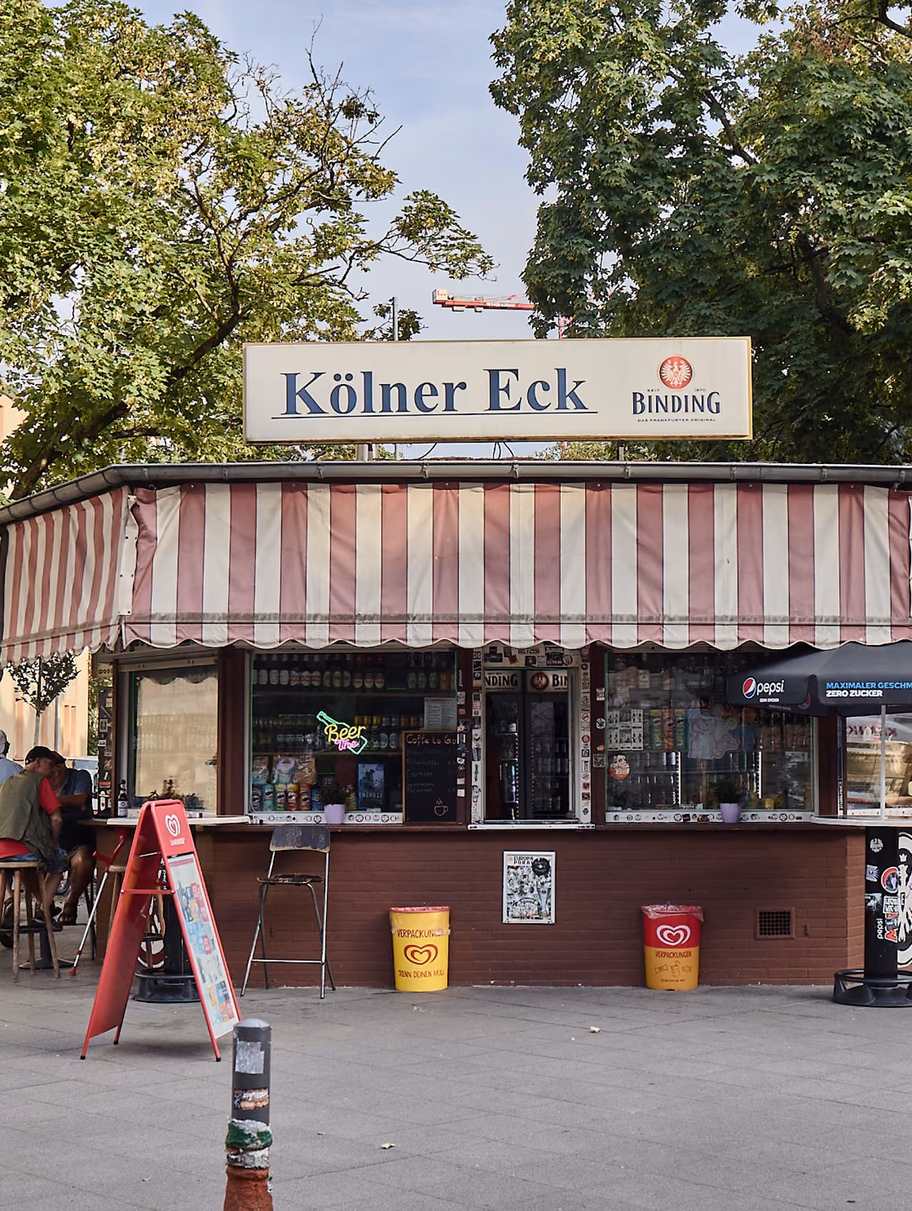Kiosk Kölner Eck mit rot-weiß gestreifter Markise unter Bäumen