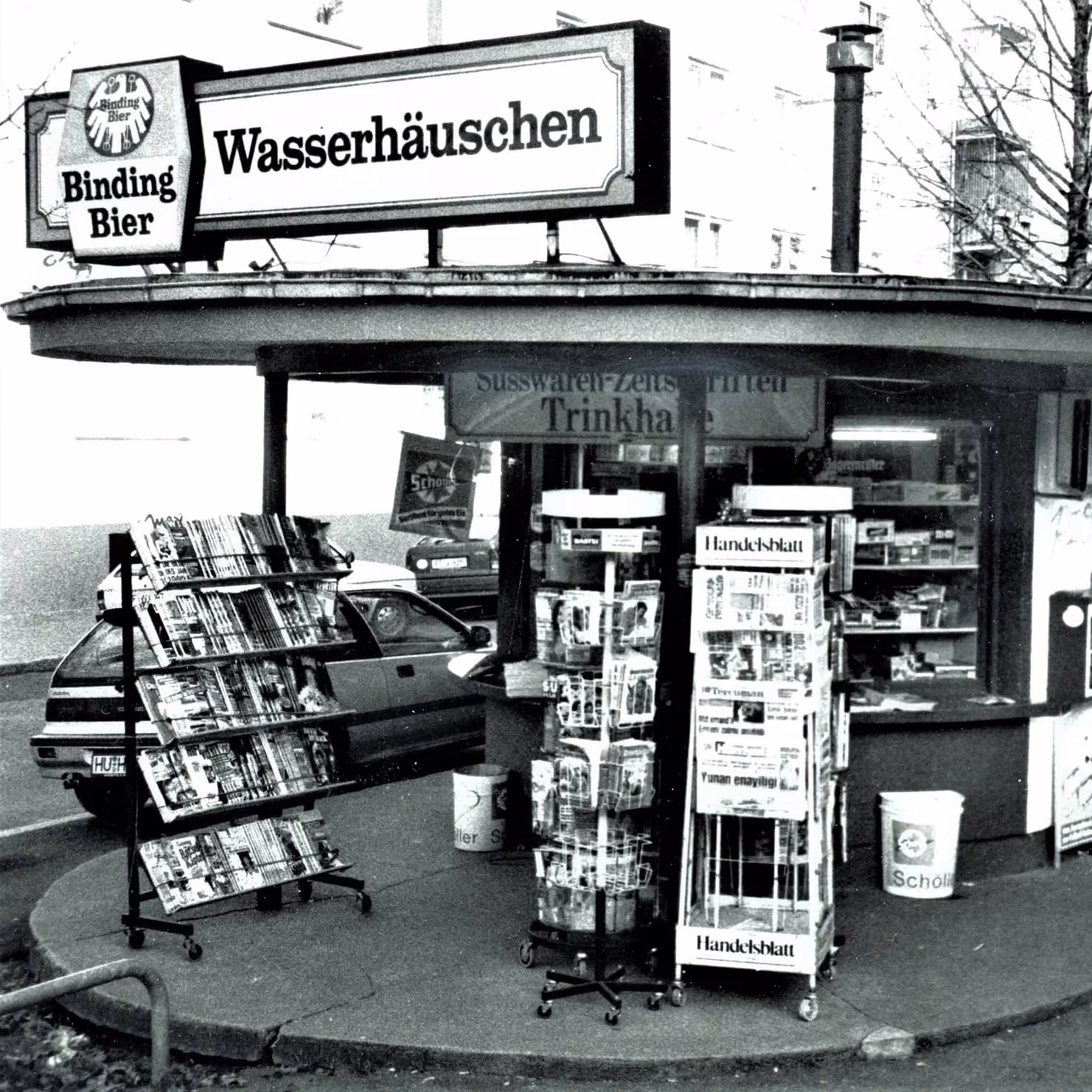 Historische Schwarz-Weiß-Aufnahme eines Frankfurter Wasserhäuschens mit Leuchtschild und Zeitungsständern davor