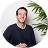 Smiling man with crossed arms standing by a green plant against a white background.