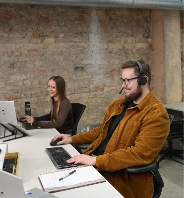 Man in headset and brown jacket typing on keyboard; woman smiling working on laptop nearby in office.