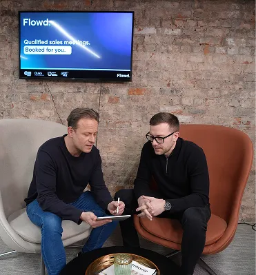 Two men seated in modern chairs discussing notes on a tablet under a screen showing Flowd sales meeting info.