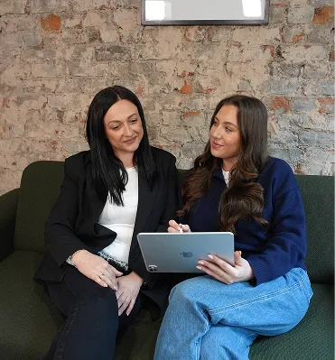 Two women sitting on a green couch, one holding a tablet while both look engaged in conversation.