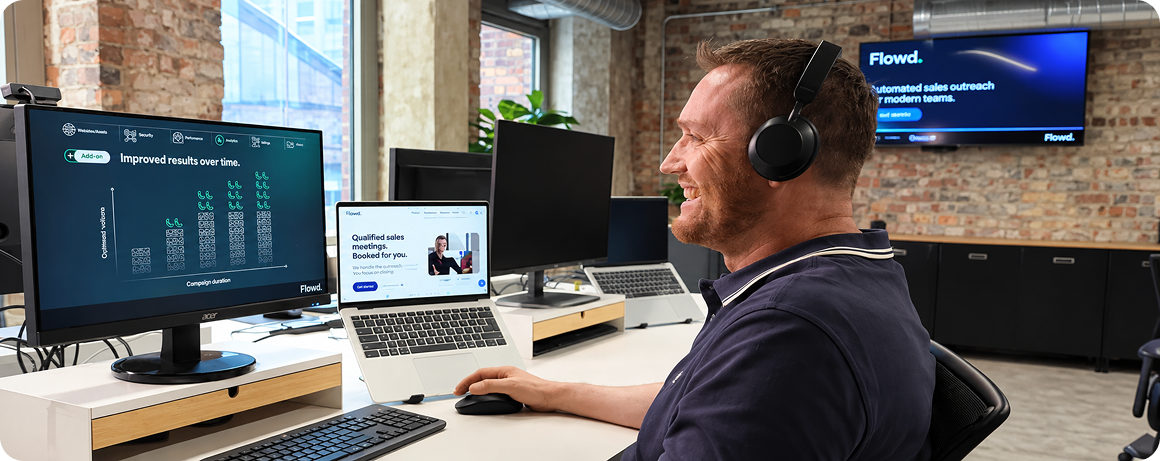 Smiling man wearing headphones works at a desk with two computer screens in a modern office.