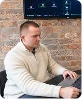 Man in a white sweater working on a laptop at a round table with a brick wall and screen behind him.