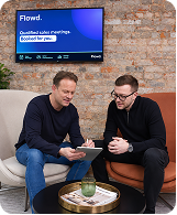 Two men sitting on sofas discussing notes in a casual office with a screen showing 'Flowed' behind them.