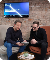 Two men sitting on sofas discussing notes in a casual office with a screen showing 'Flowed' behind them.