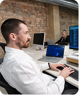 Man in white shirt working on laptop with another person blurred in office with brick wall.