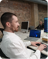 Man in white shirt working on laptop with another person blurred in office with brick wall.