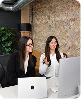 Two professional women in white and black attire engaged in conversation working at a desk with computers.
