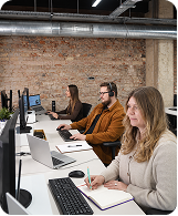 Three people working on computers at a desk in a modern office with exposed brick walls.