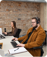 Man in brown jacket and headset working on laptop; woman in background using a phone at a desk.