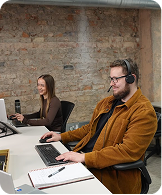 Man in brown jacket and headset working on laptop; woman in background using a phone at a desk.