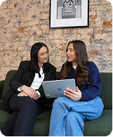 Two women sitting on a green couch, one holding a laptop, engaged in conversation.