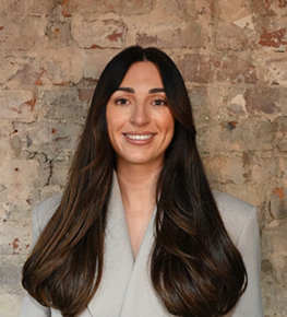 Smiling woman with long dark hair wearing a light gray blazer against a brick wall.