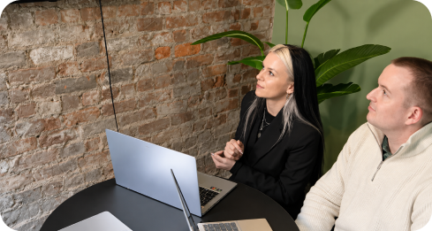 Two people sitting at a round table with laptops, looking attentively at a brick wall.