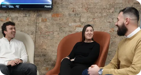 Three people smiling and sitting in modern chairs, chatting in a casual office space with a brick wall.