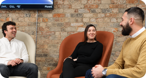 Three people smiling and sitting in modern chairs, chatting in a casual office space with a brick wall.