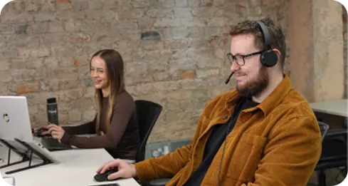 Man with headset and woman working on laptops in modern office with exposed brick wall.
