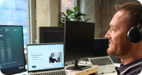 Man with headset smiling while looking at computer screens in a modern office workspace.