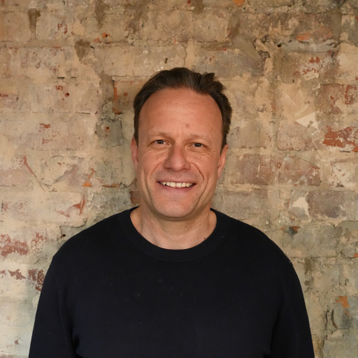 Smiling middle-aged man with short brown hair wearing a black crew neck sweater, standing in front of a weathered brick wall.