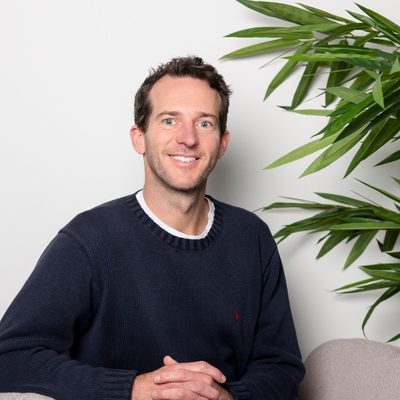 Smiling man with short curly brown hair wearing a dark blue sweater sitting indoors next to a green leafy plant.