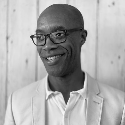Smiling man wearing glasses, a light-colored blazer, and a white shirt in front of a wooden background.