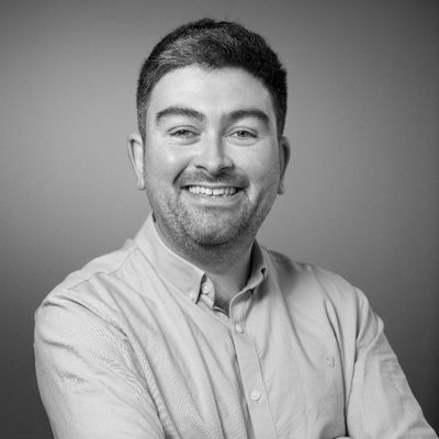Portrait of a smiling man with short dark hair and a beard, wearing a light-colored button-up shirt, in front of a plain background.