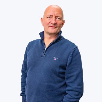 Bald man wearing a blue quarter-zip sweater with a neutral facial expression standing against a plain light background.