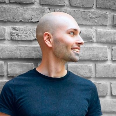 Smiling bald man in a black shirt looking to his right, standing in front of a gray brick wall.