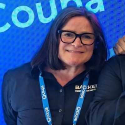 Smiling woman with shoulder-length dark hair and black glasses wearing a black shirt with a blue lanyard standing against a blue background featuring the word 'Coupa'.