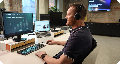 Man with headset smiling while looking at computer screens in a modern office workspace.