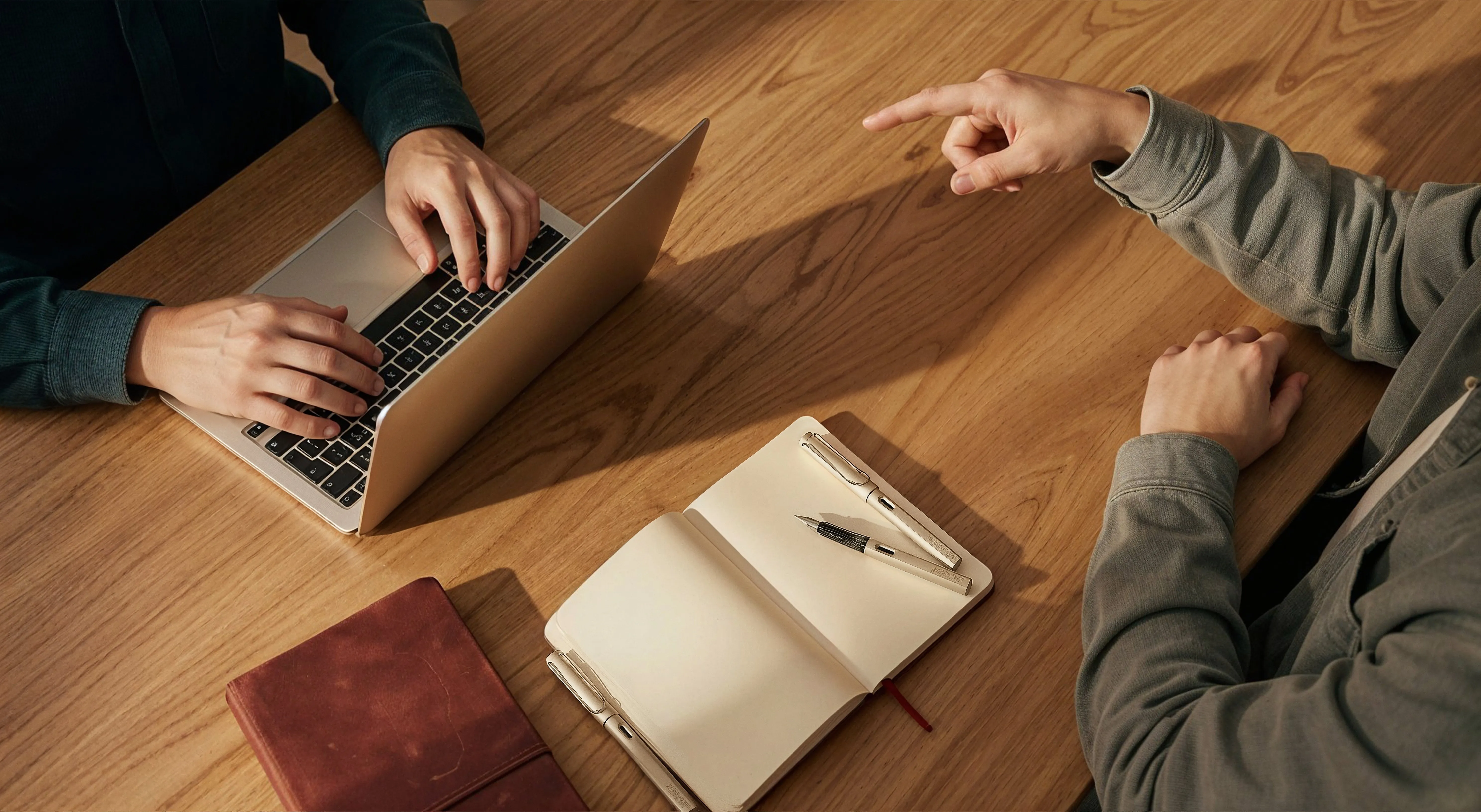 Two people at a wooden table, one typing on a laptop and the other pointing while sitting near an open blank notebook with pens.