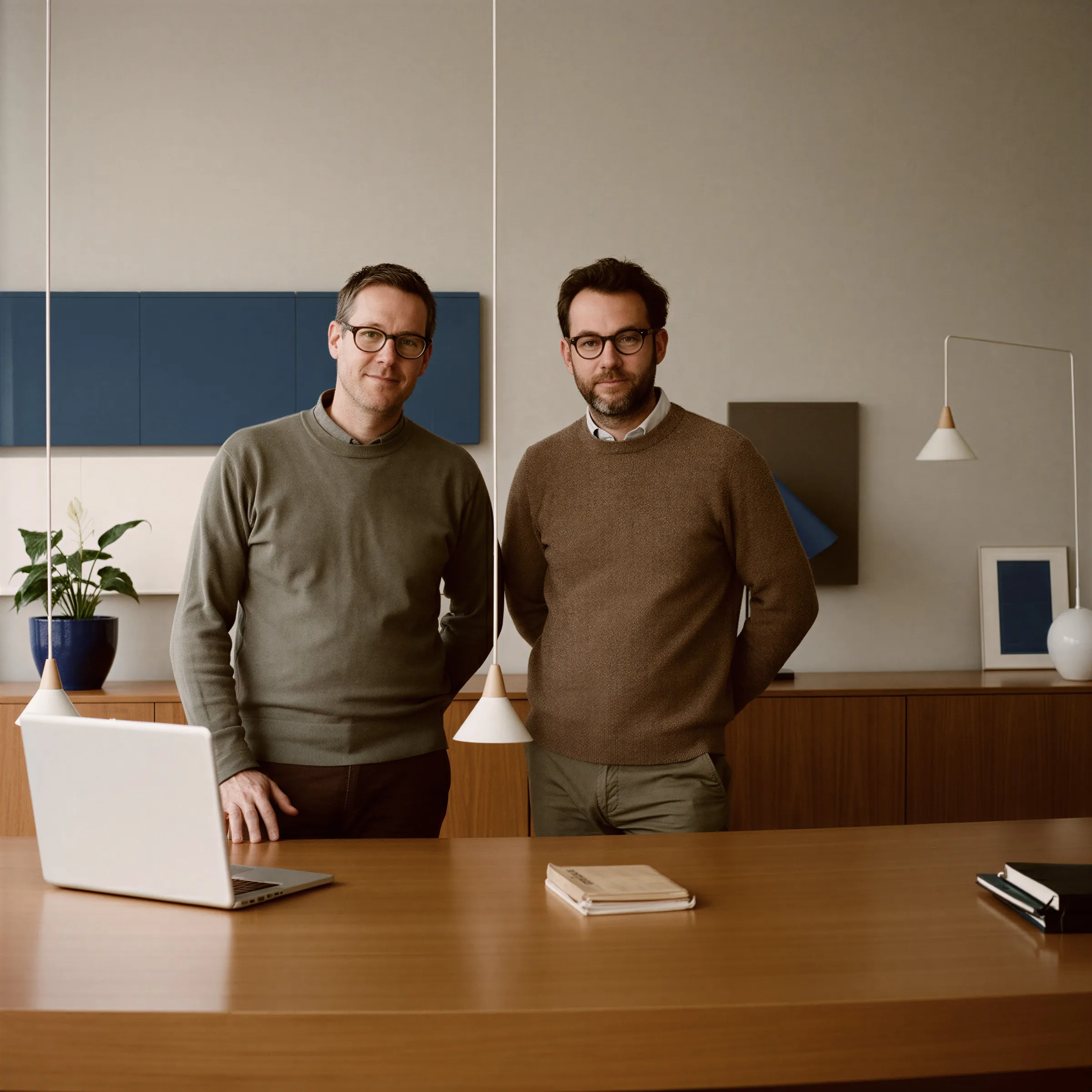 Two men wearing glasses and sweaters standing behind a wooden desk with a laptop and notebooks in a modern office.