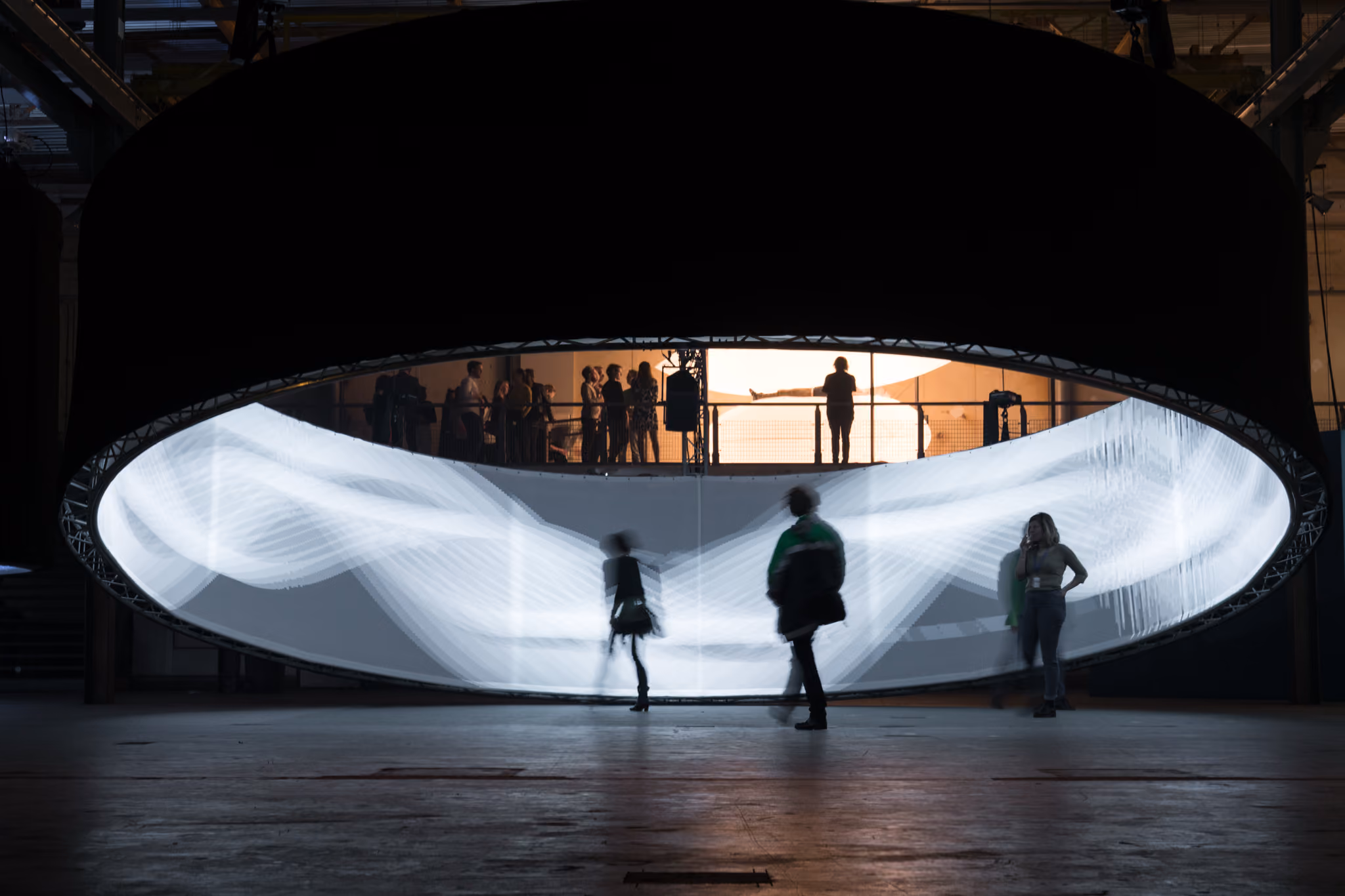 People walking in front of a large, curved light installation inside a dark industrial space.
