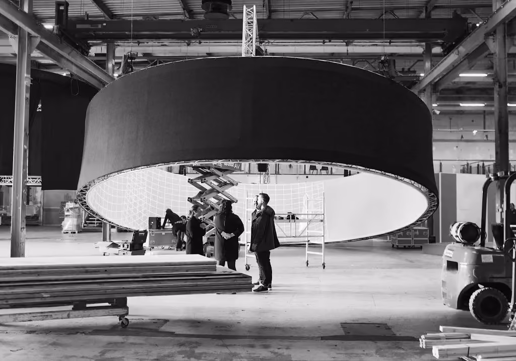 People working under a large circular structure suspended in an industrial warehouse setting.