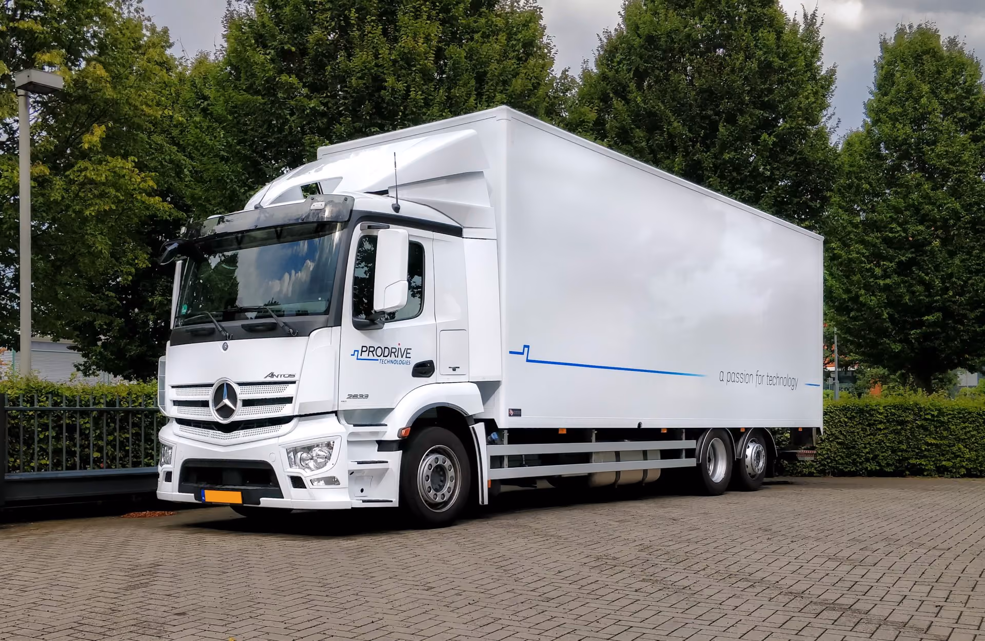 White Prodrive Technologies truck parked on a paved area surrounded by green trees under a cloudy sky.