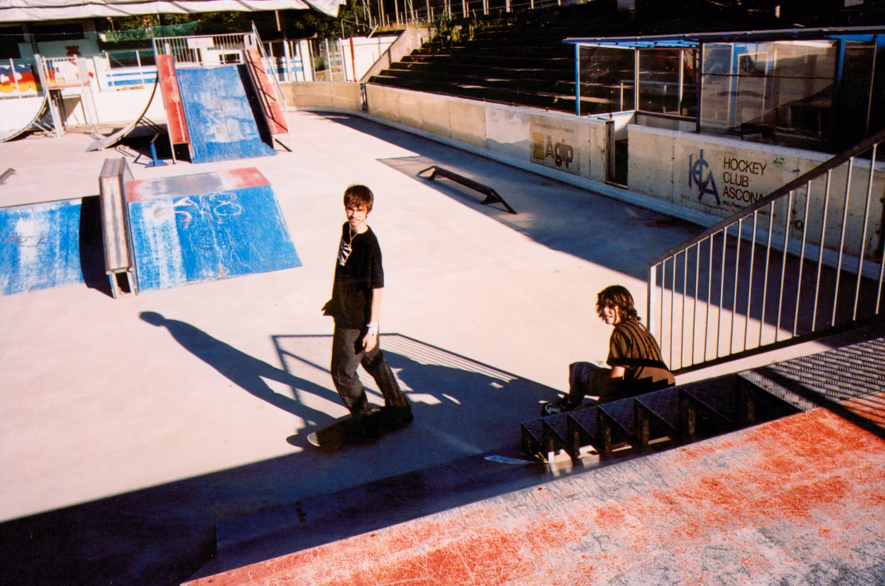 Two teenagers at a sunlit concrete skatepark, one standing on a skateboard, the other sitting on the steps