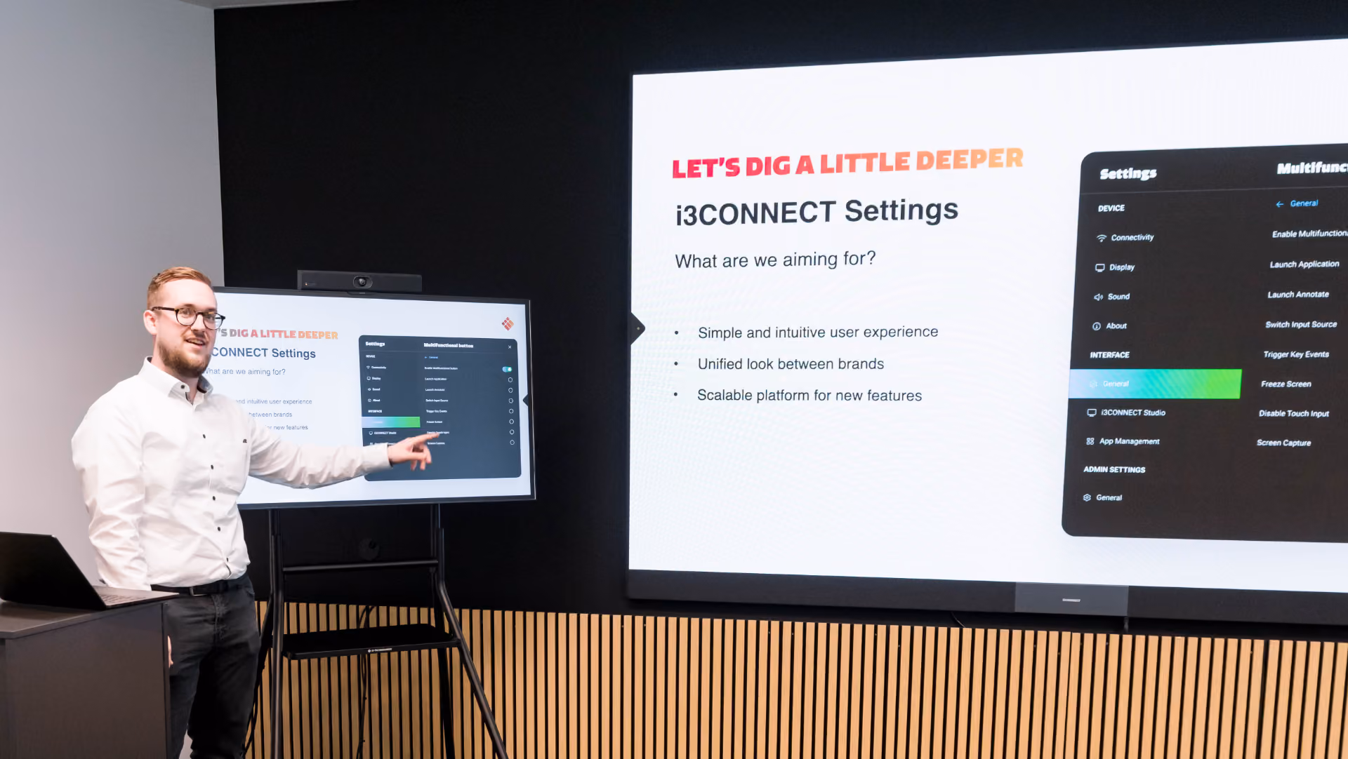 A man in glasses and a white shirt stands and points at a screen displaying i3CONNECT Settings with goals: simple user experience, unified brand look, scalable platform.
